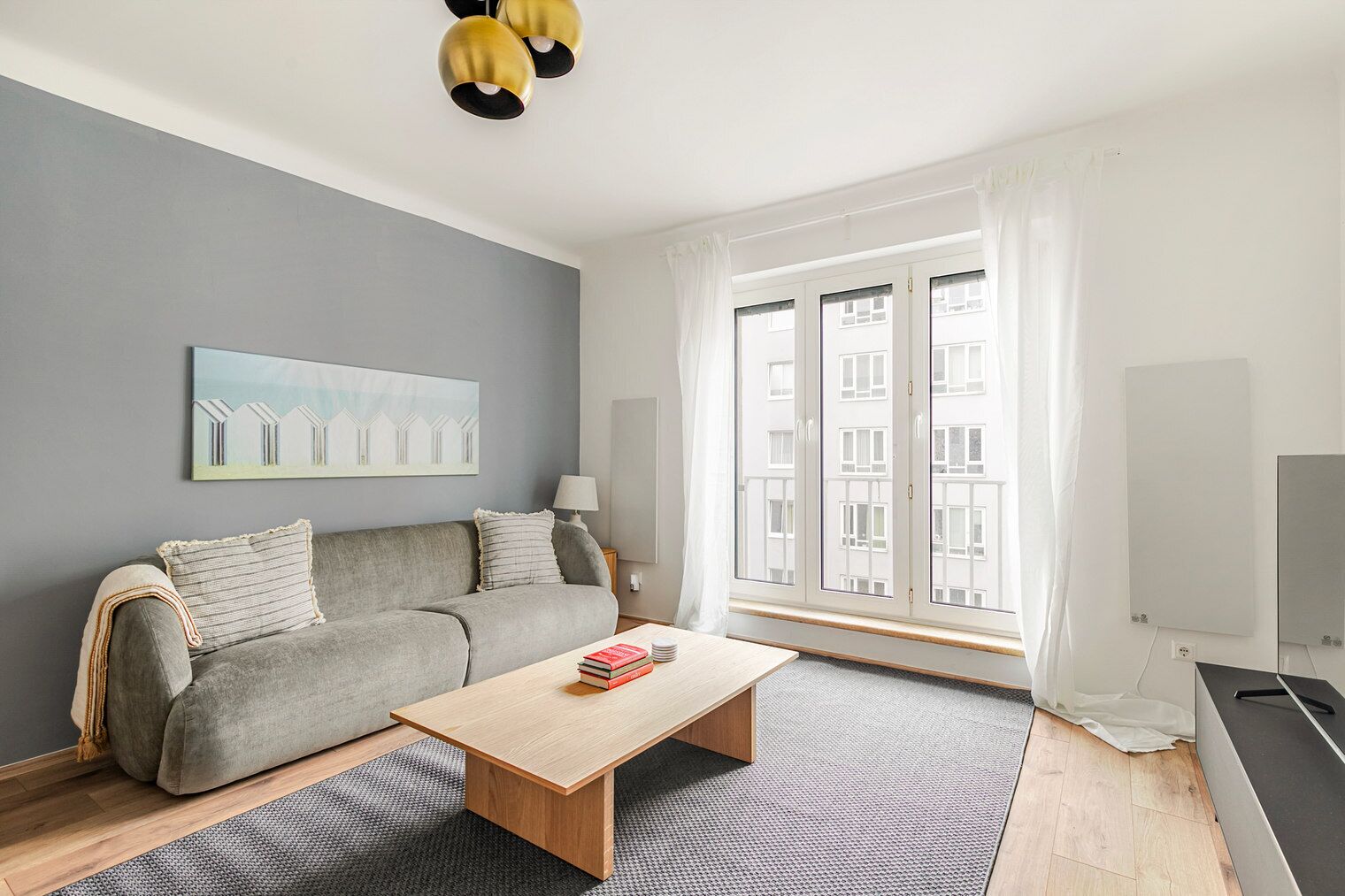 Uma sala de estar moderna com um sofá cinzento, uma mesa de centro de madeira e uma janela grande com cortinas.