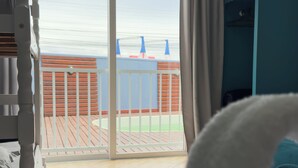 Een hotelkamer met een bed, een blauwgroene deken, een wit kussen, een witte bank, een blauwgroene garderobe, een raam met uitzicht op een balkon en de zee, en een airconditioning.