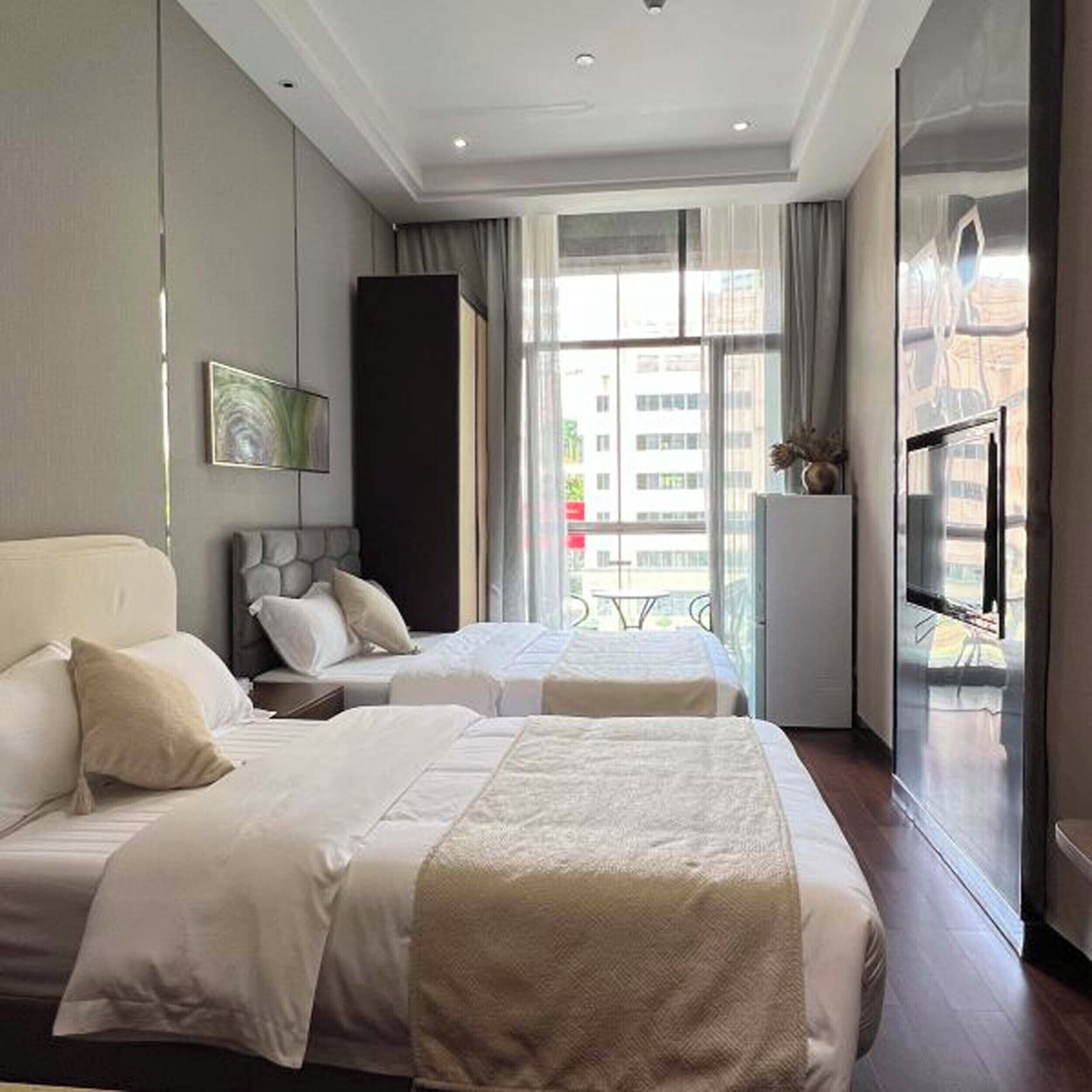 A hotel room with two beds, a large window with curtains, a flat-screen TV, and a view of buildings outside.