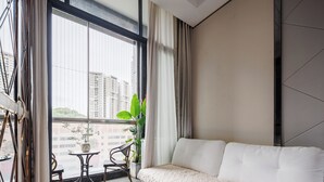 Un salon moderne avec un canapé, une petite table et un fauteuil. Il y a une grande fenêtre avec des rideaux, une plante et une vue sur les bâtiments à l’extérieur.