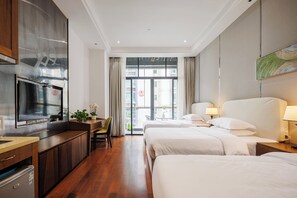 A hotel room with two beds, a desk with a TV, a small fridge, and a view of the outside balcony.