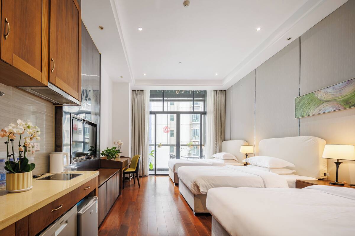 A hotel room with a kitchenette, two beds, a dining area, and a large window with curtains.