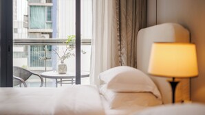 Une chambre d’hôtel avec un lit, une lampe de chevet, une petite table sur laquelle est posé un vase de fleurs, et une vue sur le paysage urbain par la fenêtre.