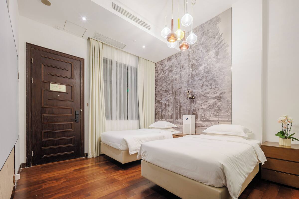 A hotel room with two beds, a wooden door, a large window with curtains, a marble wall, and a modern light fixture.