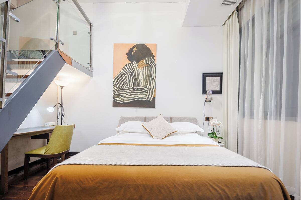 A bedroom with a bed, a desk, a chair, a staircase, and a painting on the wall.