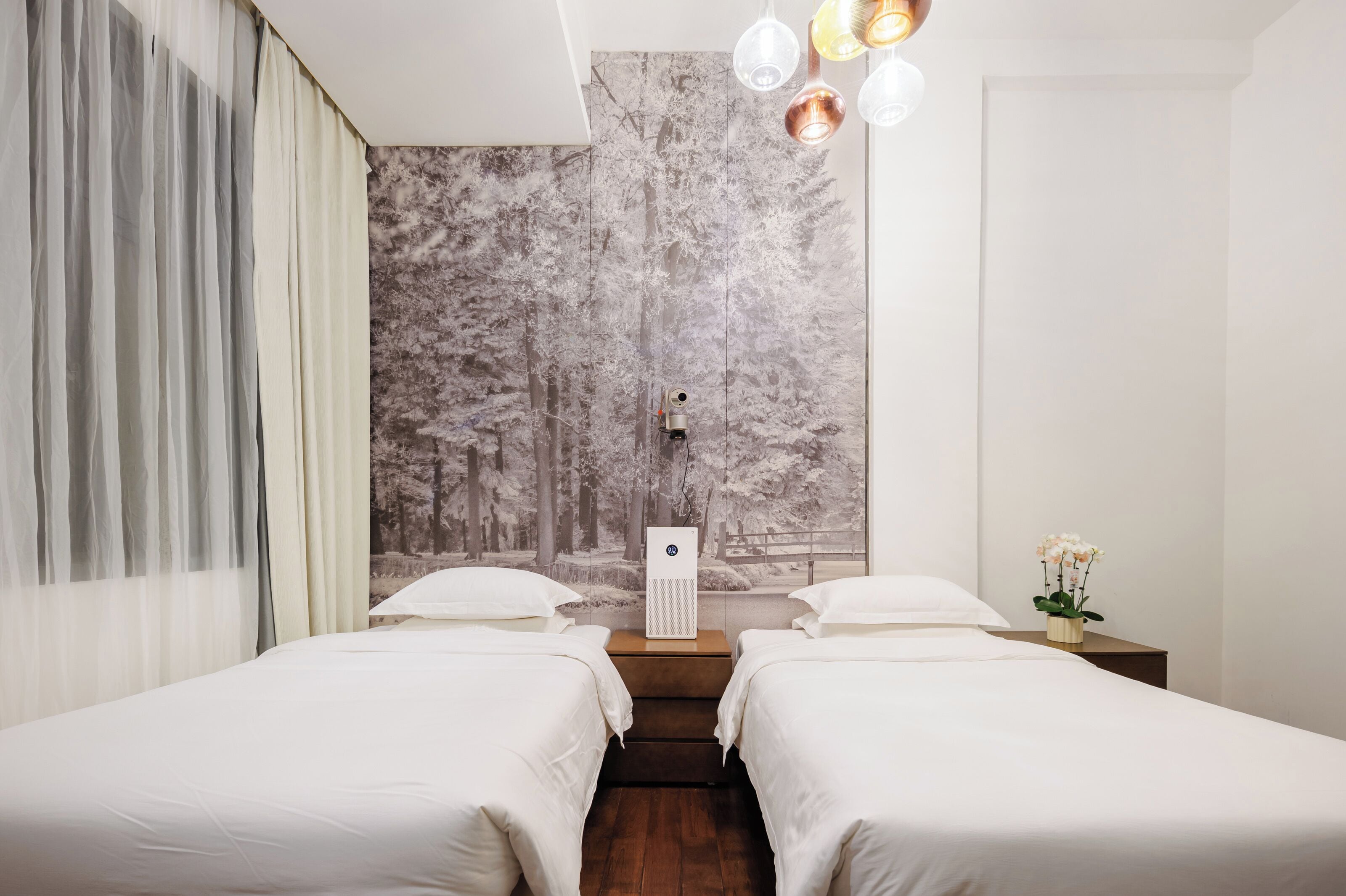 Une chambre d’hôtel avec deux lits, une grande fresque murale, une petite table sur laquelle est posé un vase de fleurs et un luminaire au plafond.