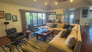 Ein geräumiges Wohnzimmer mit einer großen Couch, einem Couchtisch und einem gemusterten Teppich. Es gibt Deckenventilatoren, einen Essbereich und einen Blick ins Freie durch große Fenster.