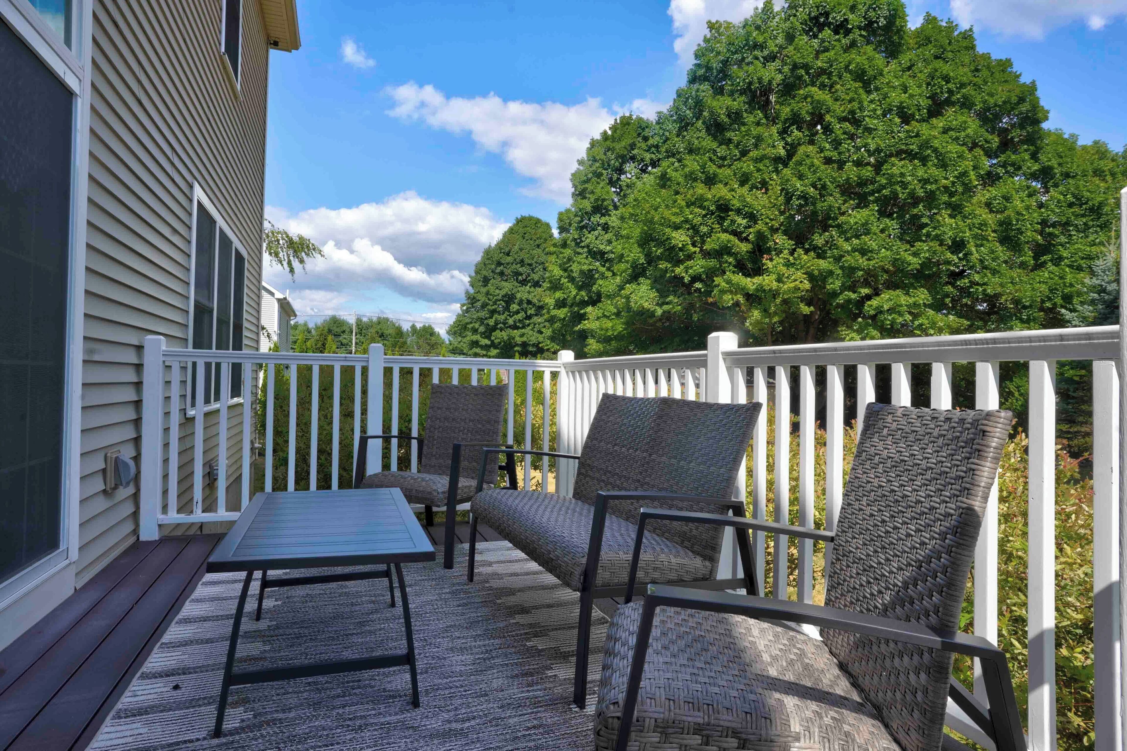 A deck with wicker furniture and a table, surrounded by a white fence, with a large tree in the background.
