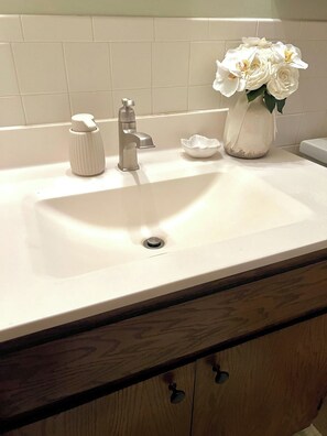 Ein Badezimmerwaschbecken mit einem modernen Wasserhahn, einem Seifenspender, einer kleinen weißen Schale und einer Vase mit weißen Blumen.