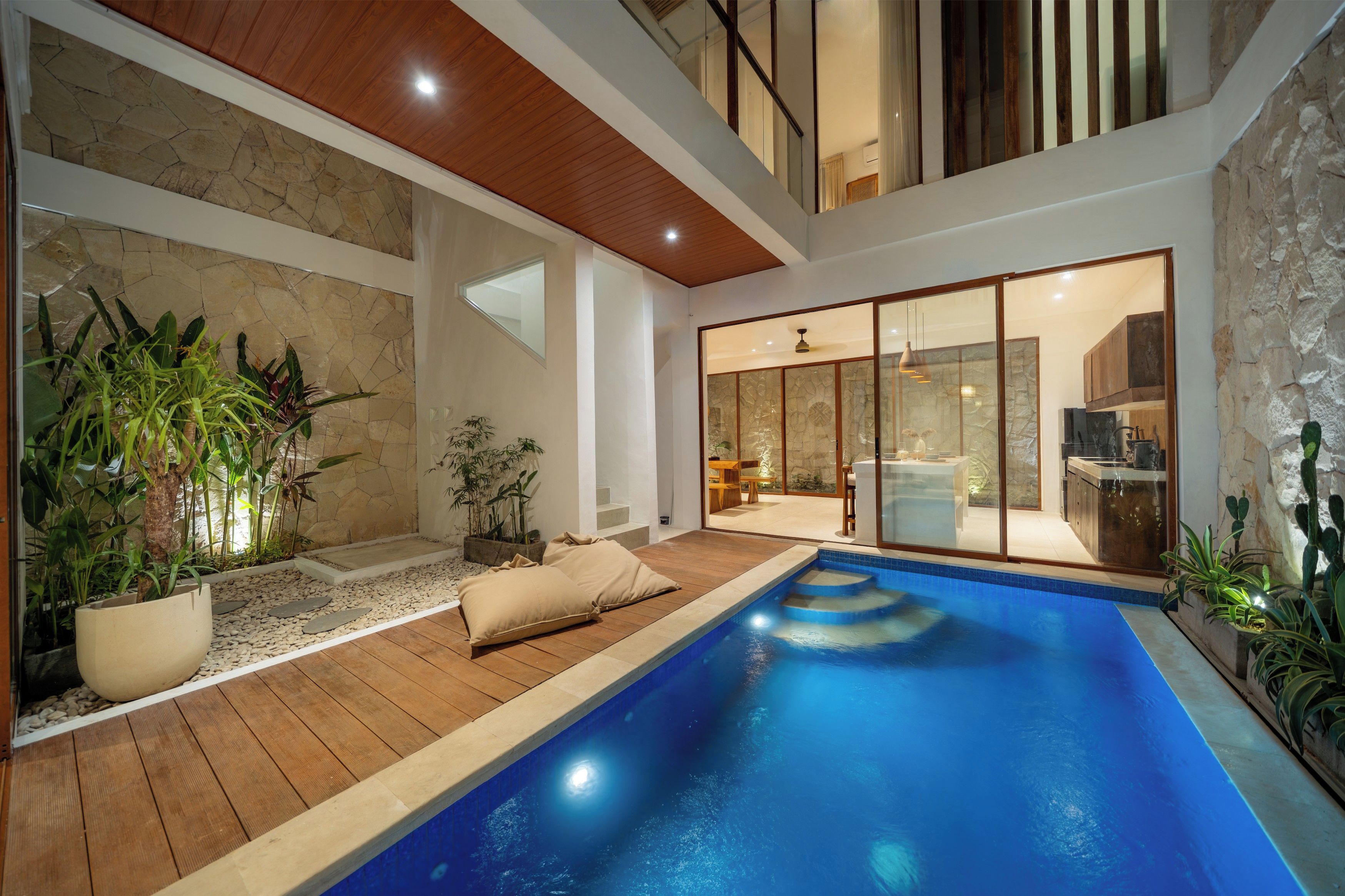A modern indoor swimming pool with a wooden deck, stone walls, and a glass-enclosed dining area.