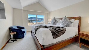 A bedroom with a large bed, a view of the outdoors, a blue chair, and a wooden nightstand. - Stunning Vista at Alpine Buzz (Frisco)