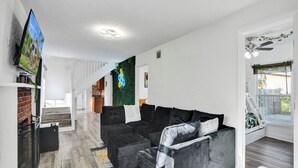 Ein modernes Wohnzimmer mit einem schwarzen Ecksofa, einem an der Wand befestigten Flachbildfernseher und einer Treppe, die in ein Obergeschoss führt.