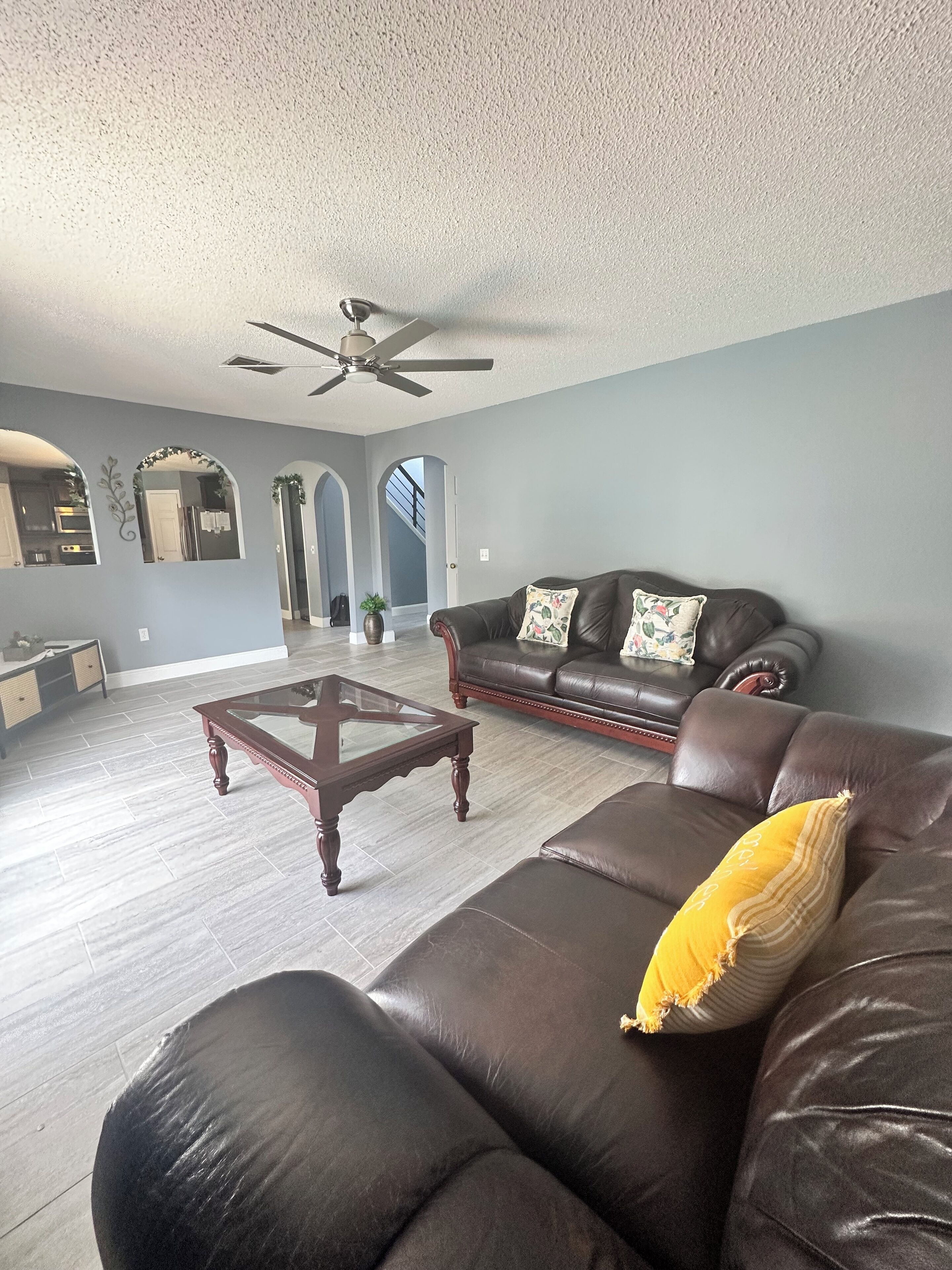 A spacious living room with a dark brown leather sofa, a glass-top coffee table, and a ceiling fan.