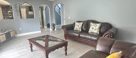 A spacious living room with a dark brown leather sofa, a glass-top coffee table, and a ceiling fan.