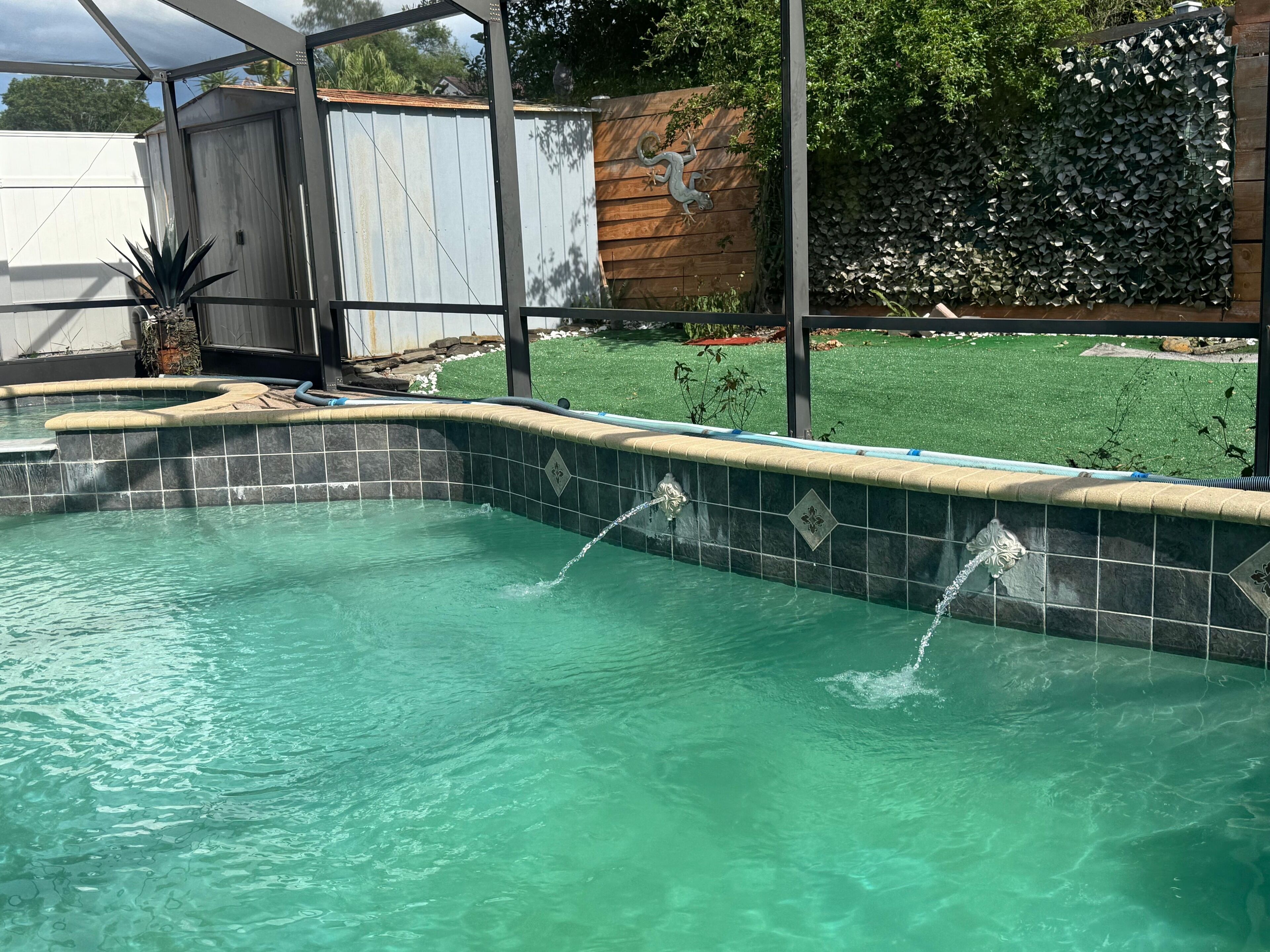 Ein Schwimmbecken mit klarem Wasser, umgeben von einer Holzterrasse und einem Metallzaun.