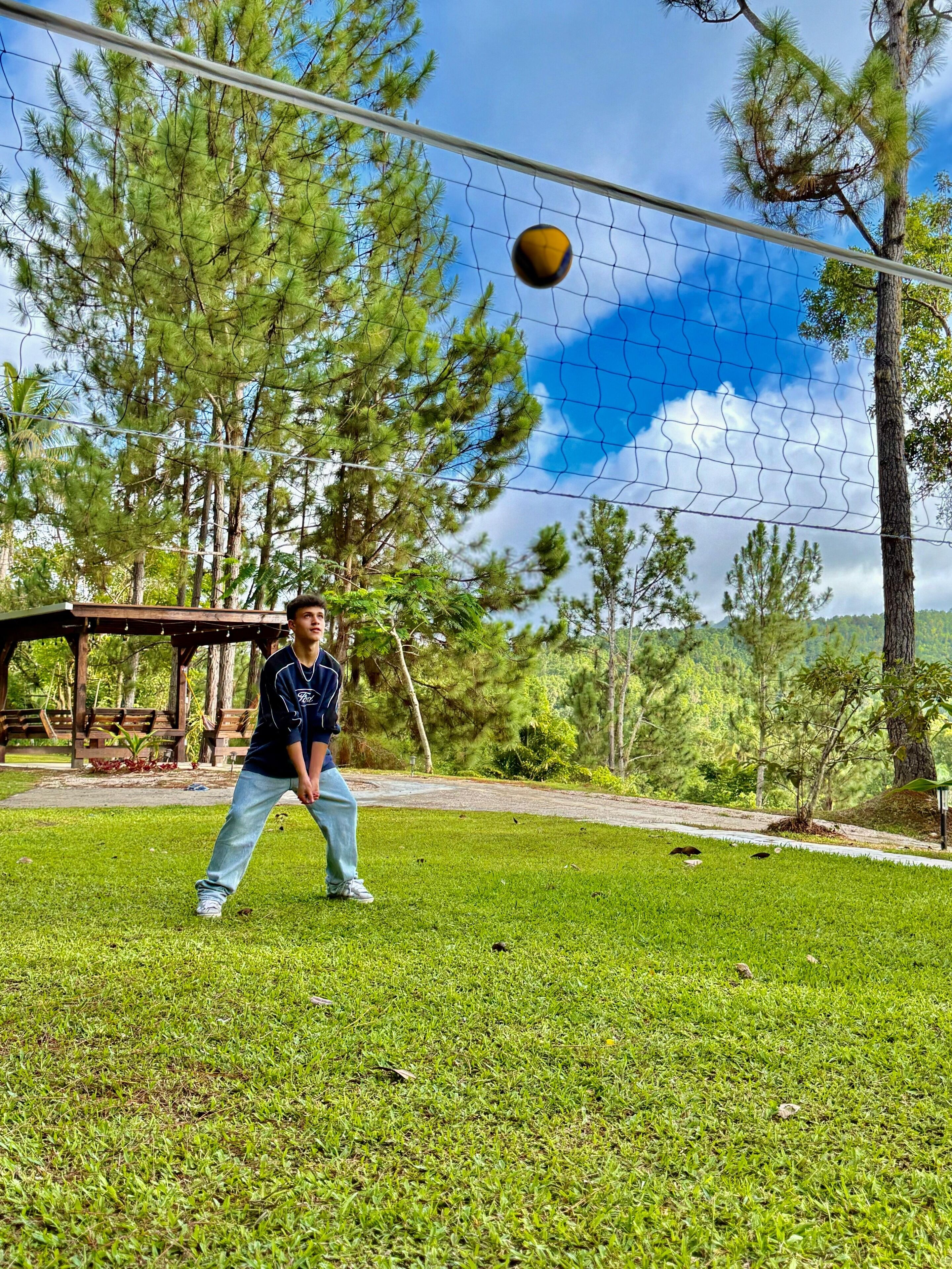 Une personne joue avec un ballon dans un espace verdoyant, avec des arbres et une structure en bois en arrière-plan.