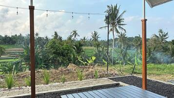 A covered outdoor seating area with a table and chairs, surrounded by a garden with palm trees and a view of rice fields.