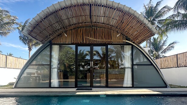 Una estructura moderna junto a la piscina, con techo curvo y ventanas amplias, rodeada por una piscina azul cristalina y palmeras.