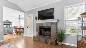 A living room with a fireplace, a television mounted above it, and a dining area with wooden chairs and a table.