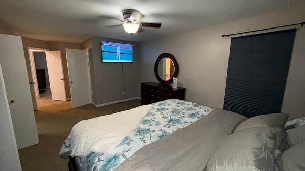 Ein Schlafzimmer mit Bett, Deckenventilator, Fernseher, Kommode und einem Fenster mit Jalousien.