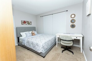 Ein Schlafzimmer mit Bett, Schreibtisch und Stuhl. Es gibt ein Fenster mit Jalousien, eine Wanddekoration und eine Tür.