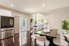 A modern kitchen with stainless steel appliances, a dining area with a dark wood table and cushioned chairs, and a living area with a sofa and a fireplace. - The Royal Estate (Fort Washington)