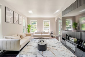 A modern living room with a sofa, a coffee table, and a TV. There are two windows with plants and a view of greenery outside. - The Royal Estate (Fort Washington)