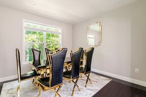 A dining room with a round table and six chairs, a large window with blinds, and a wall-mounted mirror.
