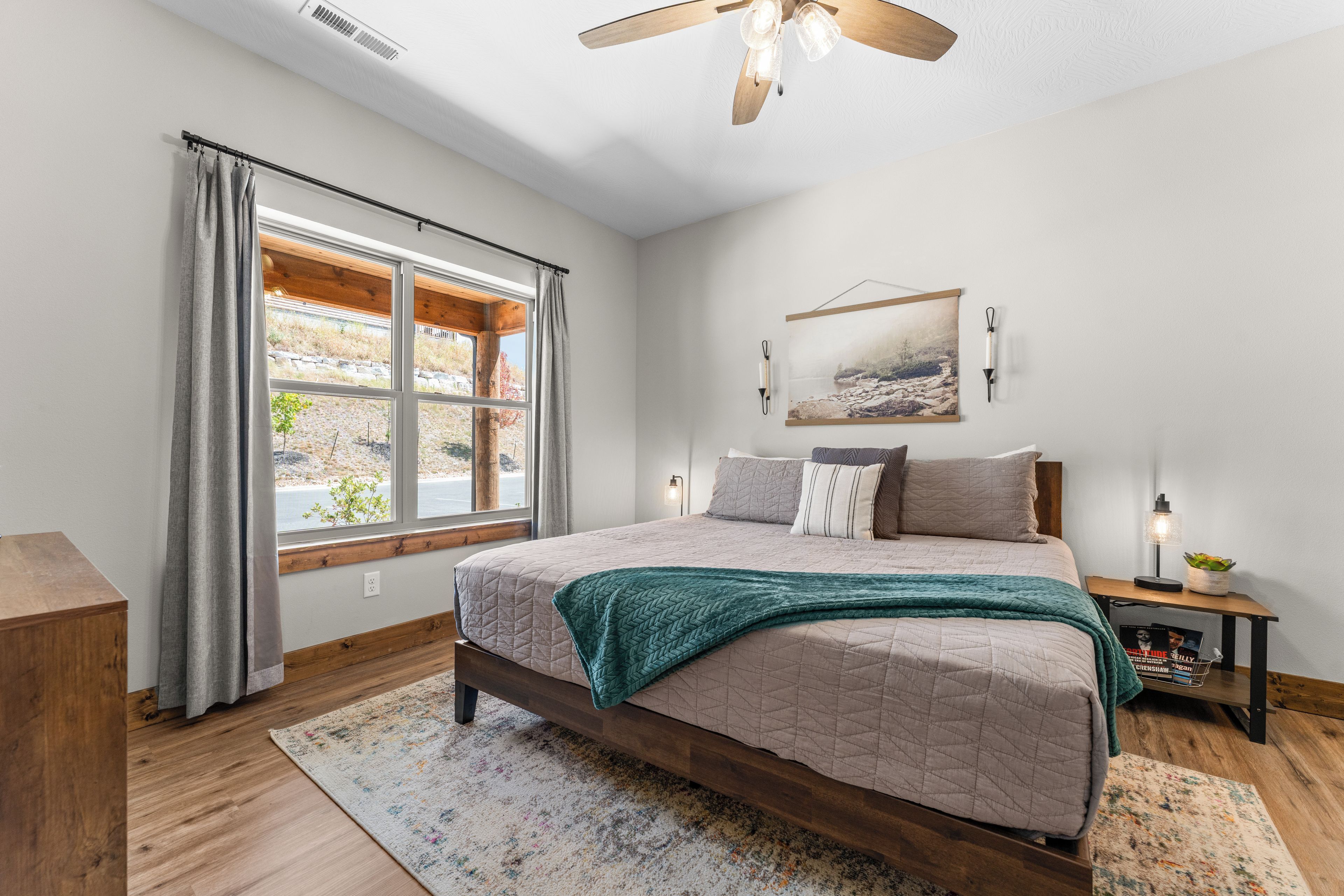 A bedroom with a large bed, a window with curtains, a ceiling fan, a bedside table with a lamp, and a wall-mounted picture.