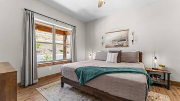 A bedroom with a large bed, a window with curtains, a ceiling fan, a bedside table with a lamp, and a wall-mounted picture.