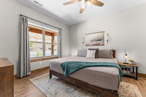 A bedroom with a large bed, a window with curtains, a ceiling fan, a bedside table with a lamp, and a wall-mounted picture.
