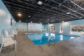 Una piscina interior con piso azul, sillas reclinables blancas y techo con vigas a la vista.