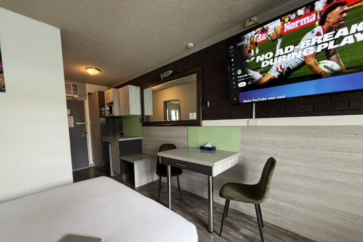 A hotel room with a bed, a desk, a chair, a TV mounted on the wall, and a kitchenette area.