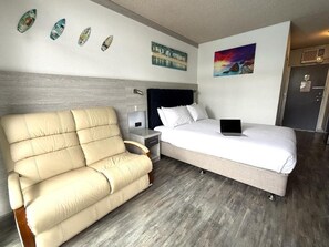 A hotel room with a bed, a sofa, a nightstand, a laptop on the bed, and wall art. - Le George Motel (Port Macquarie)