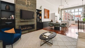 A modern living room with a fireplace, a television, and a dining area with a view of the city. - The Higham (Fremantle)