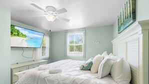 A bedroom with a bed, a ceiling fan, a TV, a window, and a painting on the wall.