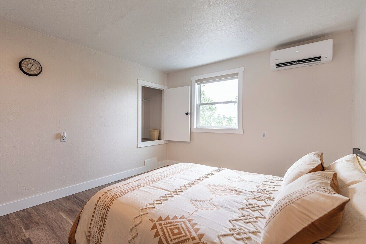A bedroom with a bed, a window, an air conditioning unit, and a clock on the wall.