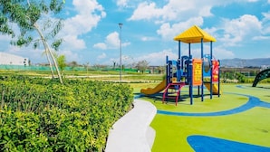 A playground with a yellow slide and climbing structures, surrounded by green hedges and a clear sky.