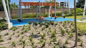 An outdoor exercise area with exercise equipment, a shaded canopy, and a building in the background.