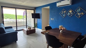 A modern living room with a blue accent wall, a dining area with a table and chairs, a sofa, a flat-screen TV, and an air conditioning unit.