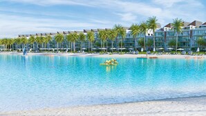 A resort pool with clear blue water, surrounded by palm trees and a row of hotel buildings.