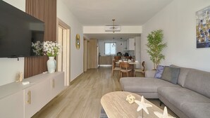 A modern living room with a flat-screen TV, a sofa, a dining area, and a kitchen in the background.