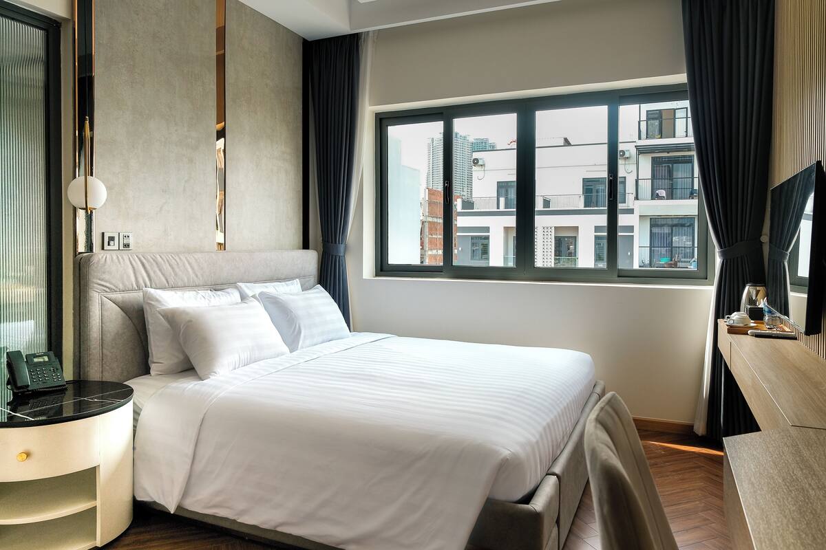 Ein Hotelzimmer mit einem großen Bett, einem Nachttisch mit Lampe, einem Schreibtisch mit Stuhl und einem Fenster mit Blick auf die Stadt.
