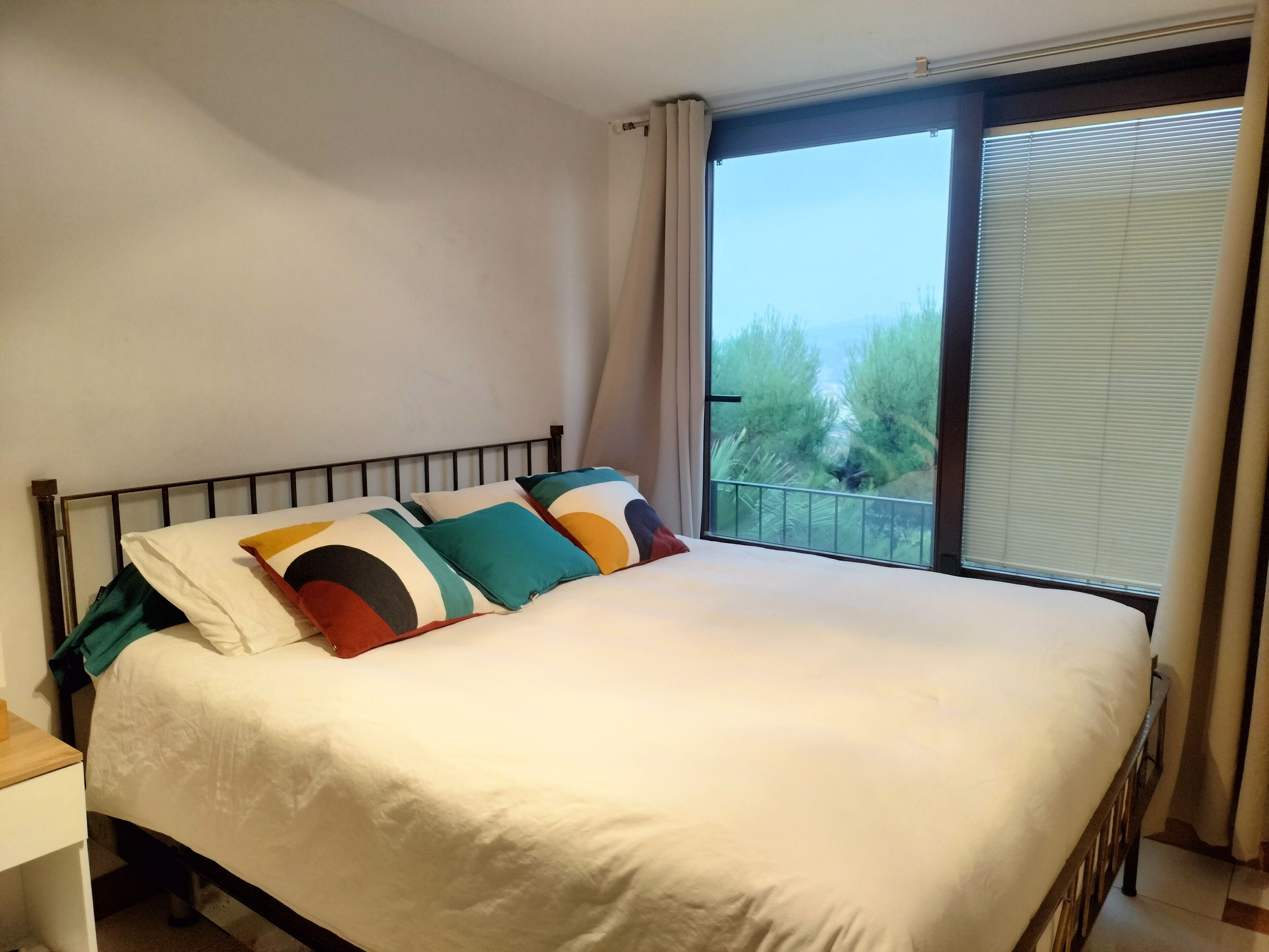 A bedroom with a large bed, a nightstand, and a window with a view of greenery.