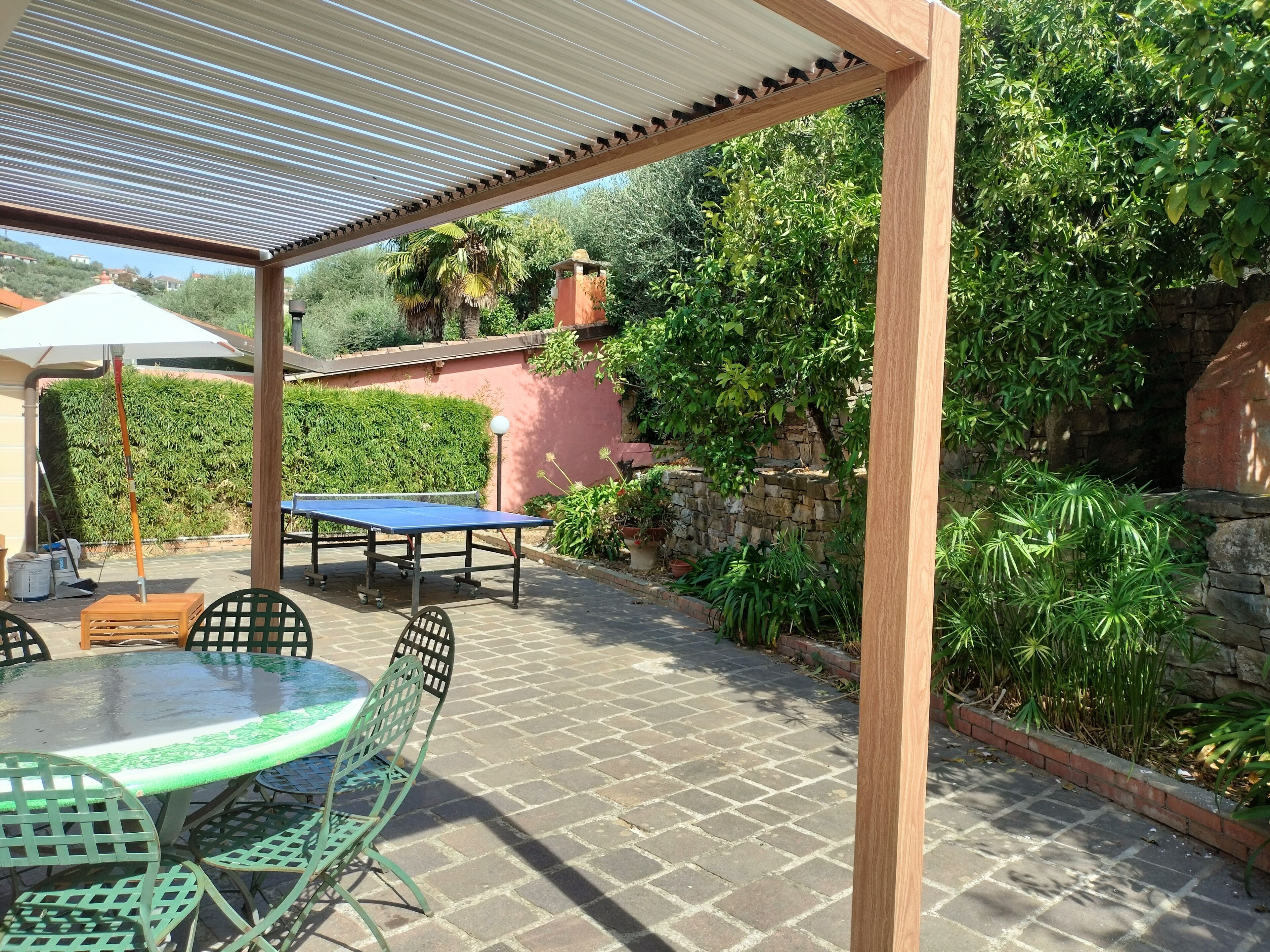 An outdoor patio with a table and chairs, a pergola overhead, and a stone wall with greenery.
