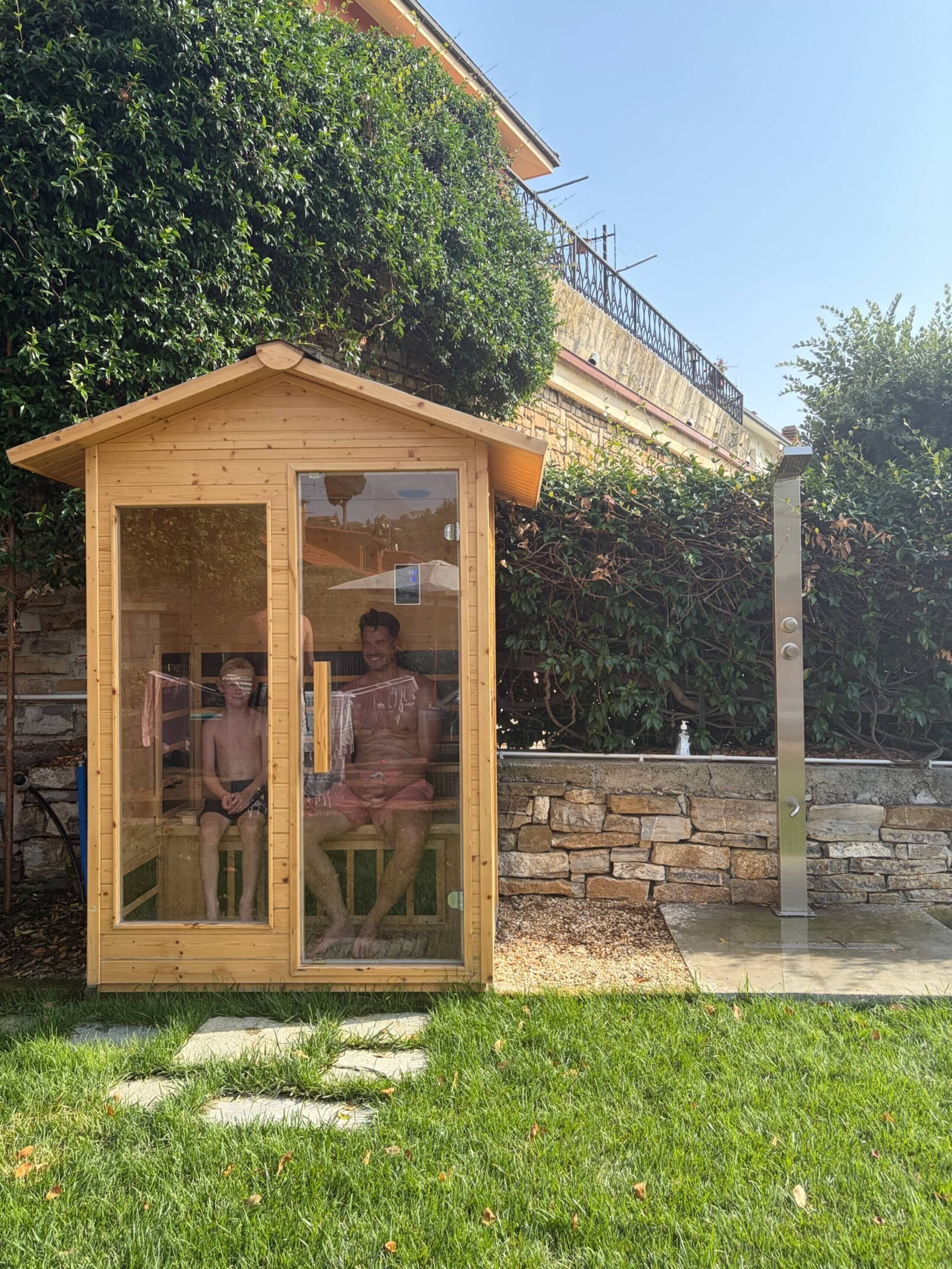 Une structure de sauna en bois dotée d’une porte en verre, située dans un jardin avec un mur de pierres et de la verdure en arrière-plan.