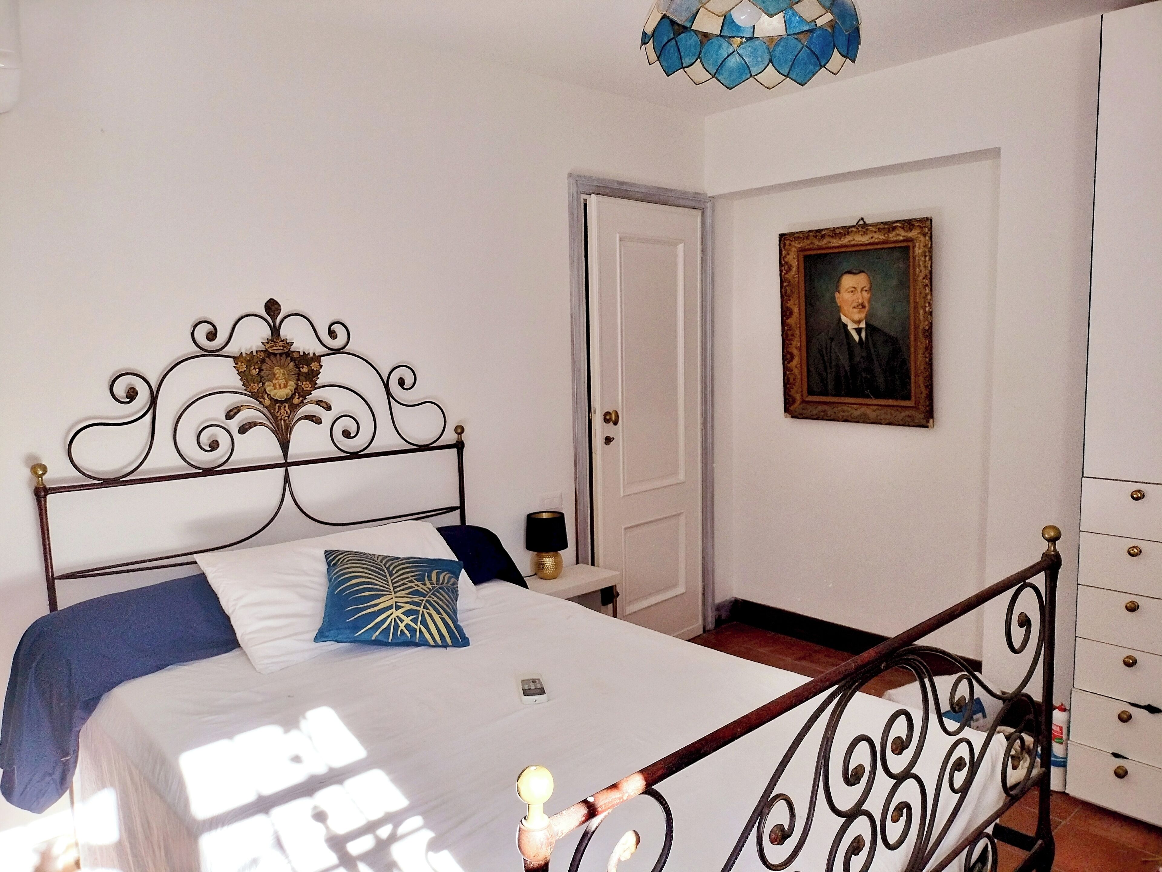 A bedroom with a bed, a bedside table, a lamp, a dresser, a framed portrait, and a door.