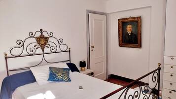 A bedroom with a bed, a bedside table, a lamp, a dresser, a framed portrait, and a door.