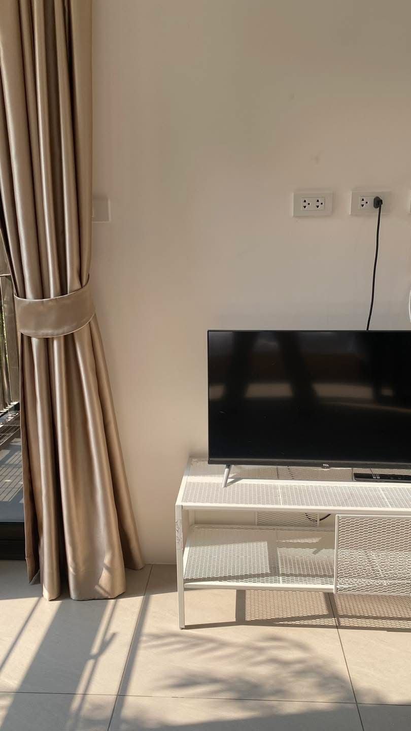 A modern living room with a flat-screen TV on a white stand, beige curtains, and a tiled floor.