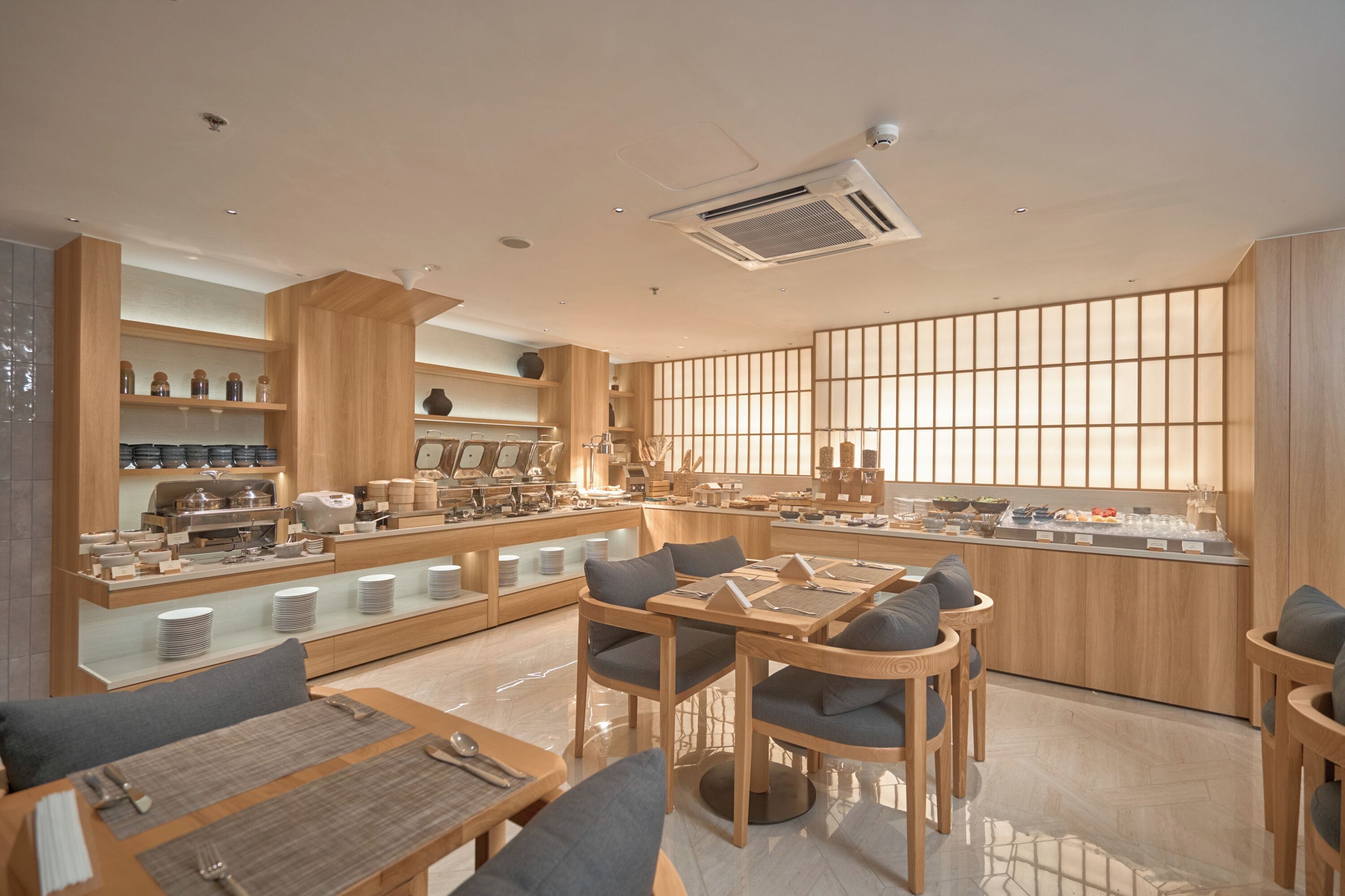 Una cafetería moderna y bien iluminada, con mobiliario de madera, una barra central y un gran ventanal con persianas verticales.